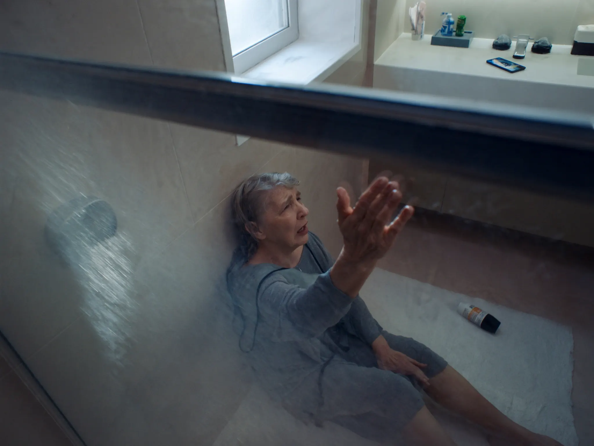 An older woman sits on the bathroom floor after a fall, reaching up