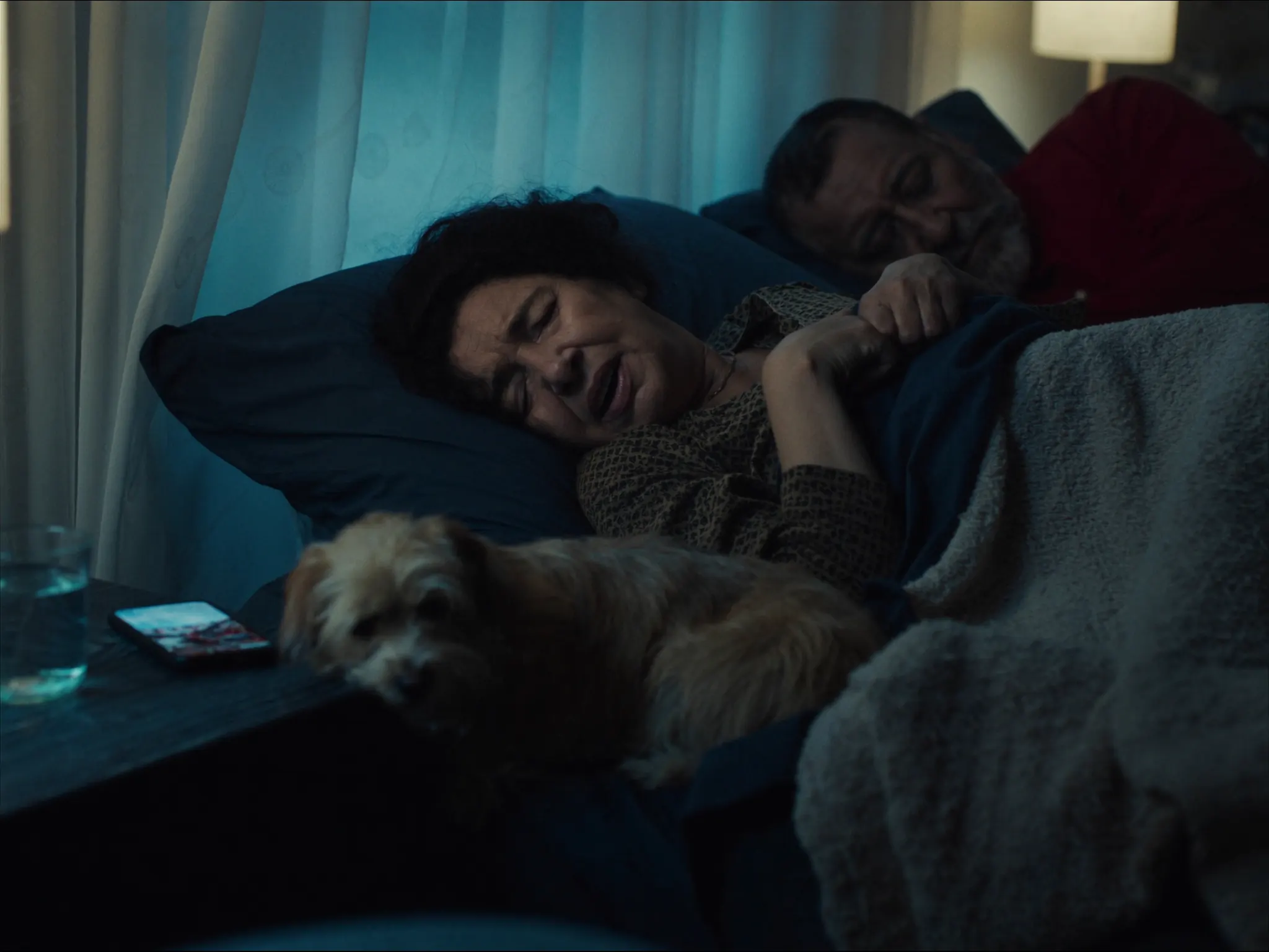 An older woman lies awake and unwell in bed at night beside her sleeping partner and dog